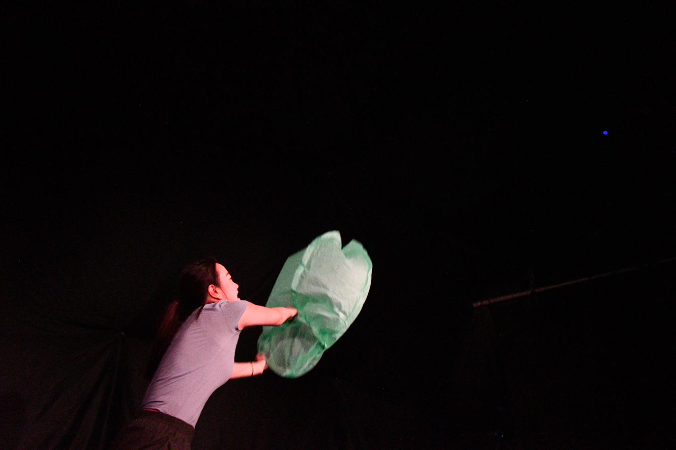 A performer releases a glowing green lantern into darkness in Between Strands.