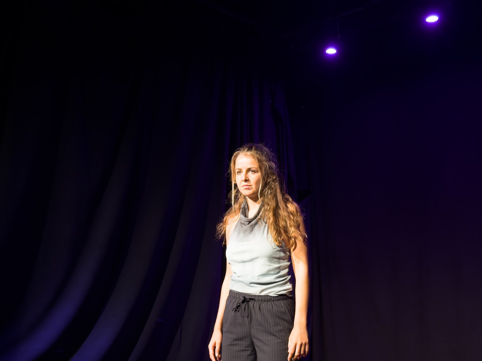 Lalia stands alone in a pool of light against a dark curtain, looking out with a quiet, serious expression.