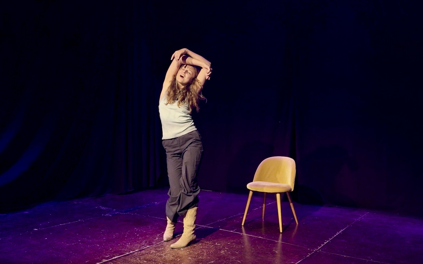 Lalia stretches both arms over her head, eyes closed, beside a yellow chair on a purple-lit stage.
