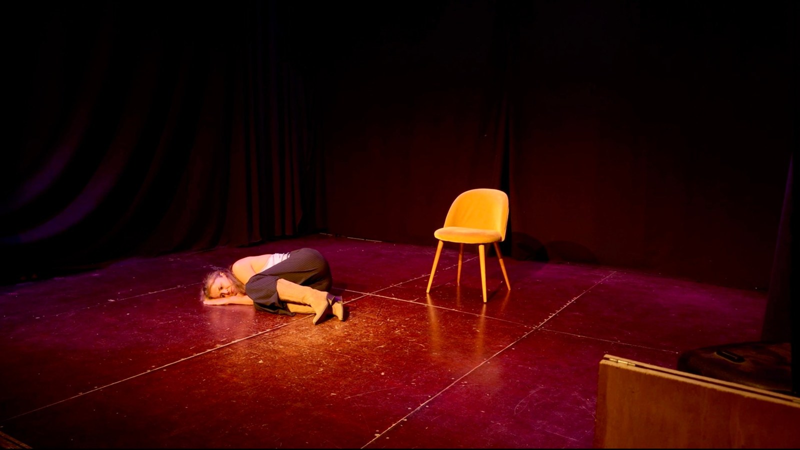 Lalia curled up on the stage floor, resting her head on her arm, with an empty yellow chair nearby.