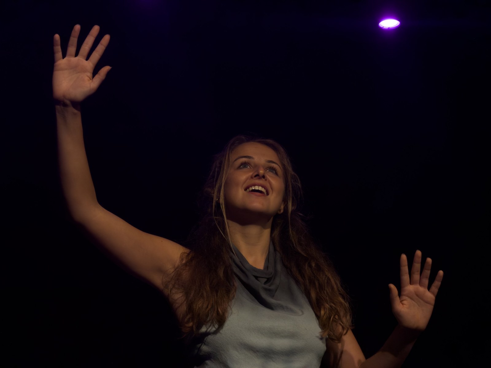 Lalia gazes upward with a smile, both hands raised, lit warmly against black.