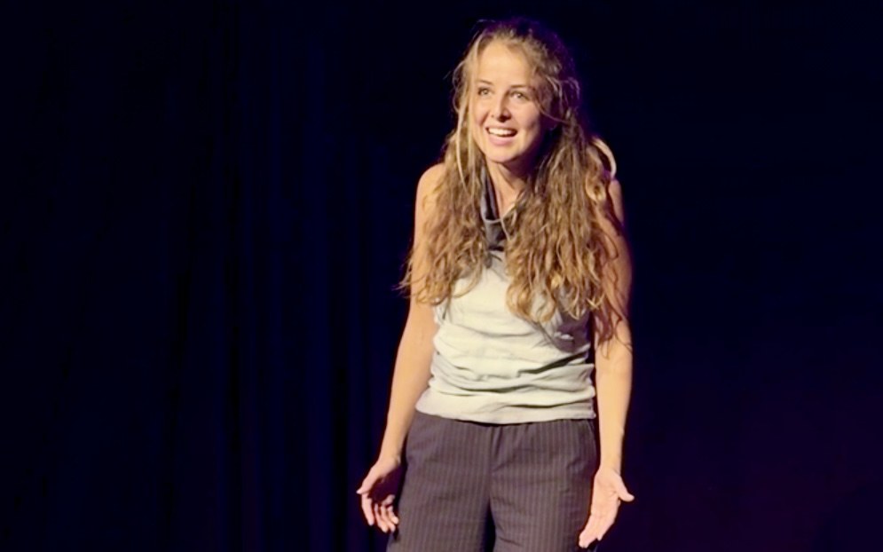 Lalia laughs, caught mid-gesture with open hands, in front of a dark curtain.