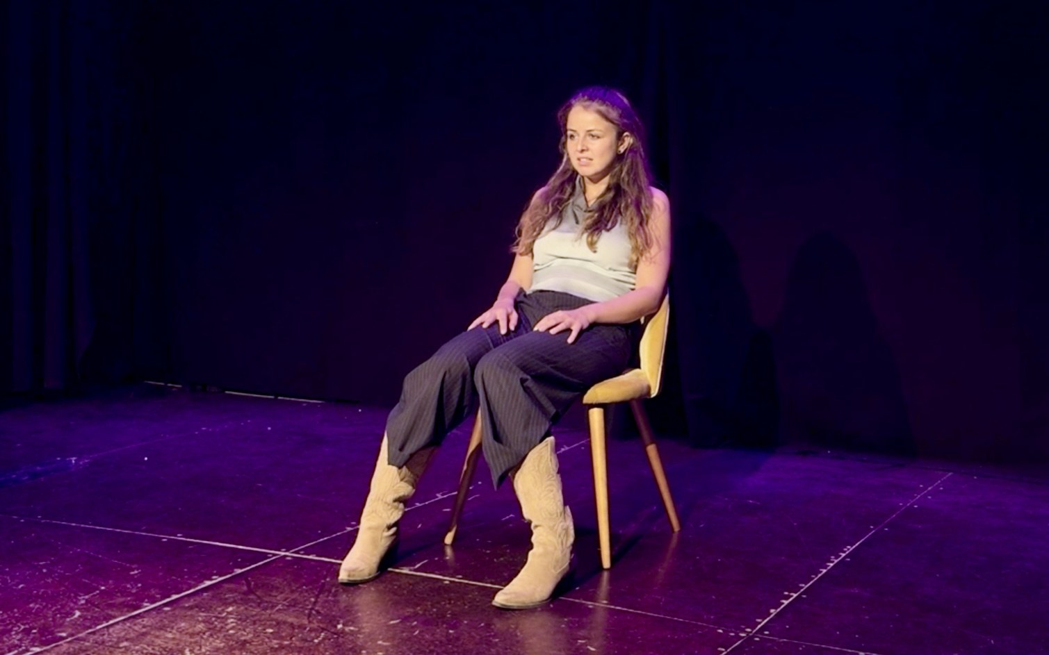 Lalia sits on a wooden chair, hands on her knees, in cowboy boots and pinstripe trousers.