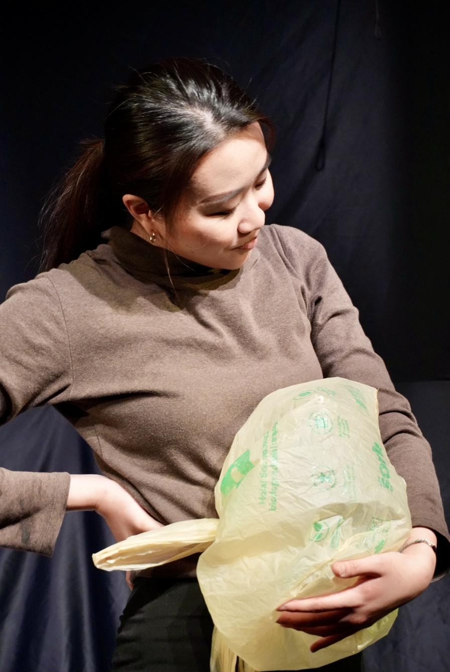 A performer cradles a plastic bag like something precious, lit against black.