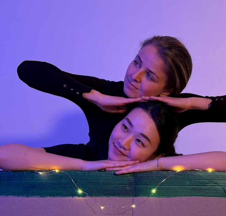 The two performers of Out of the Bin, one resting her chin on the other's hands above a fairy-lit cardboard box.