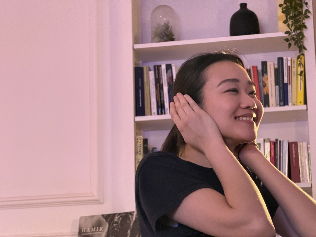A performer smiling, hands to her ears, in front of a bookshelf.
