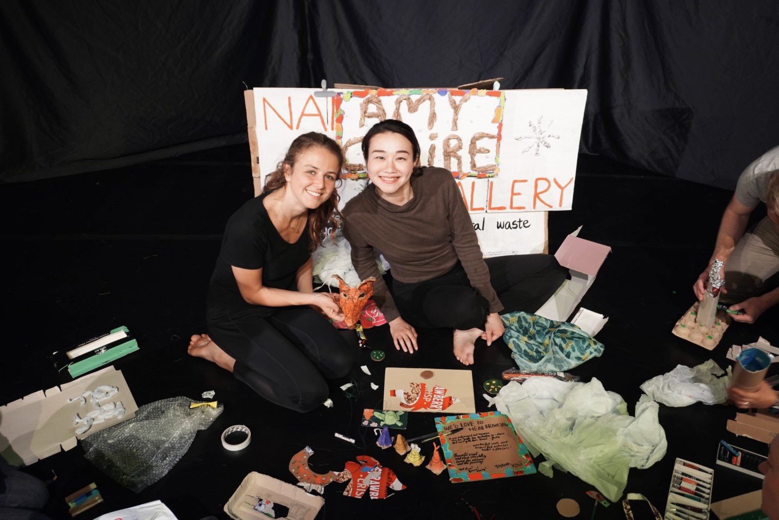 Two performers sit on the floor smiling beside a hand-made fox mask and a cardboard gallery sign.