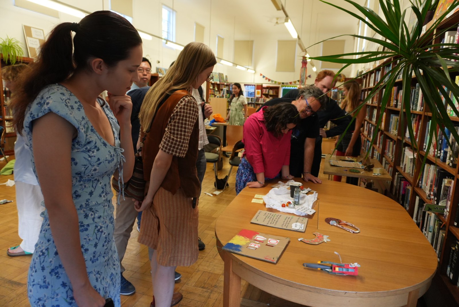 Workshop participants gather around a table, looking at objects made from waste in a library.