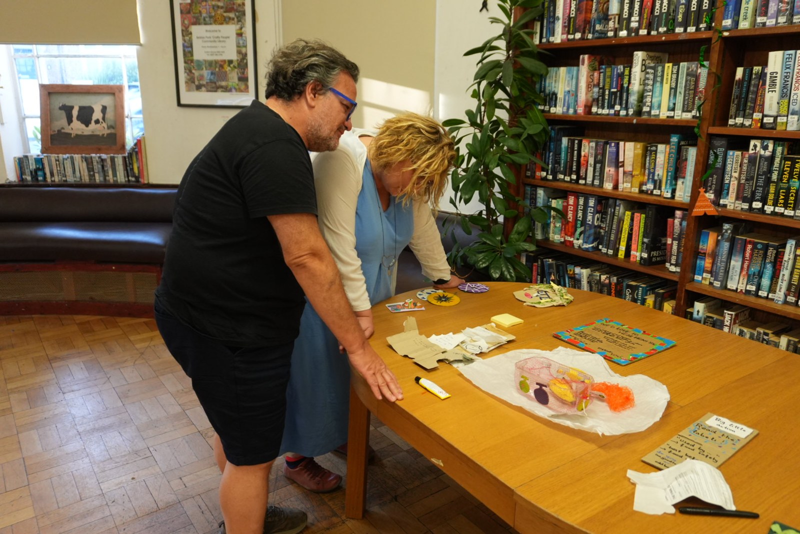 Two participants look closely at art objects made from waste on a library table.