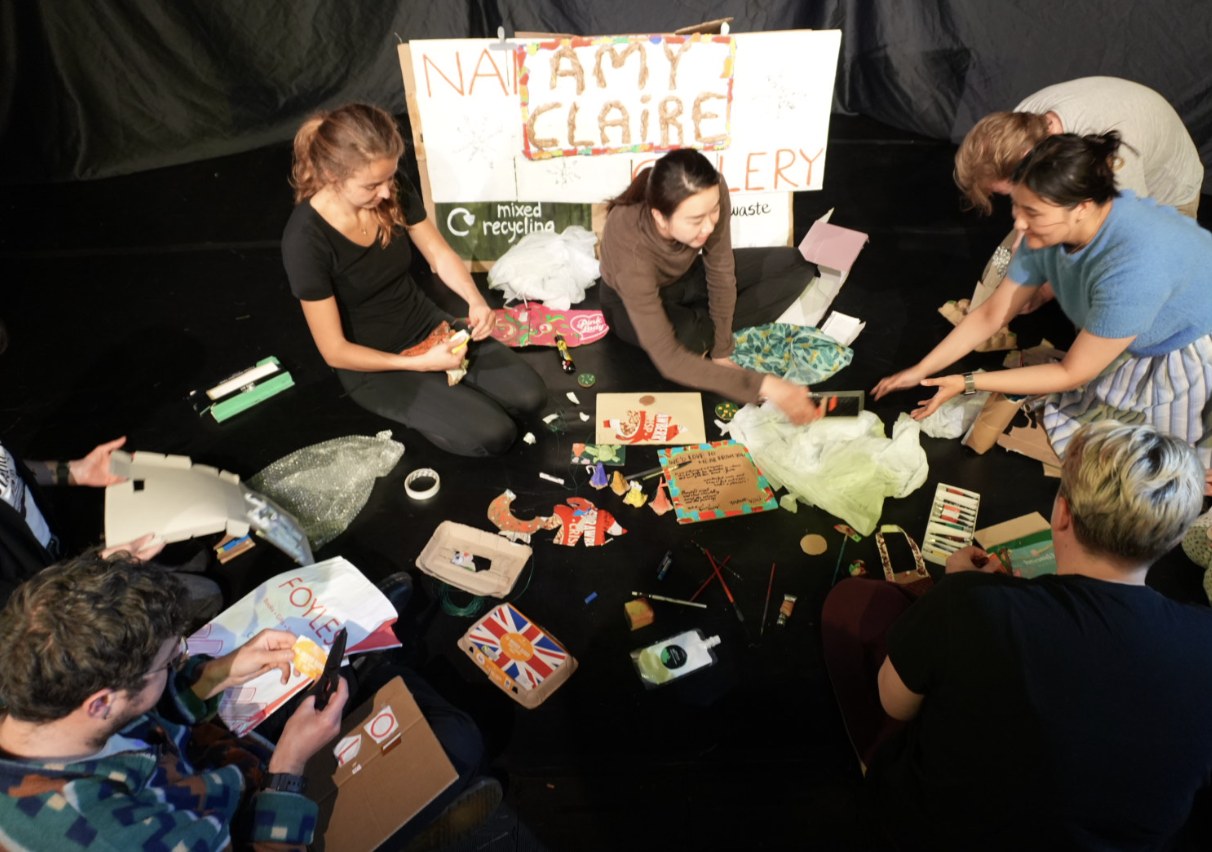 A circle of participants making art from cardboard and waste on a dark stage.